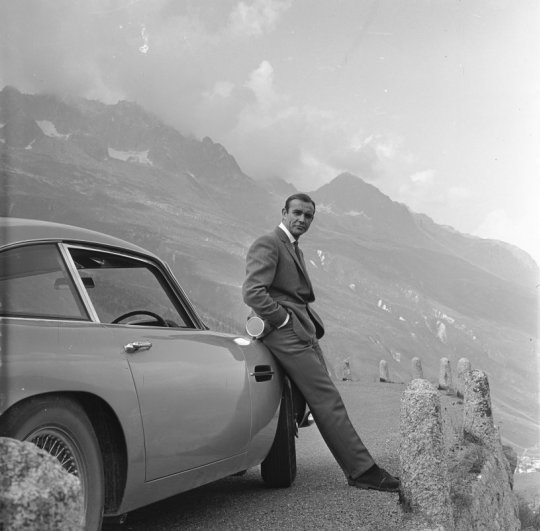 . SEAN CONNERY relaxes on the bumper of his Aston Martin DB5 during the filming of location scenes for 'Goldfinger' in the Swiss Alps. Copyright Notice - © 1964 Danjaq, LLC and United Artists Corporation. All rights reserved.